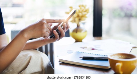 Close-up Young Asian Woman Touching On Mobile Screen To Order Food While She Work At Home With Notebook And Coffee Drink On Desk