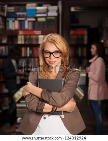 close-up of young attractive caucasian girl with geeky eyeglasses holding a book in her arms with bookshelves and other people in background