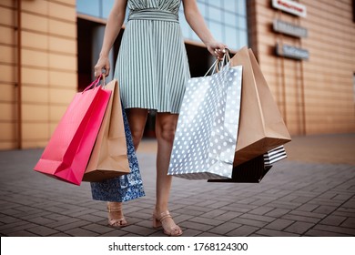 Closeup Of Young Attractive Woman Holding A Few Shopping Bags With Newly Purchased Goods And Clothes. Girl Holds Colourful Packs Full Of Bought Things In Her Hands. Shopping And Spendings Concept.