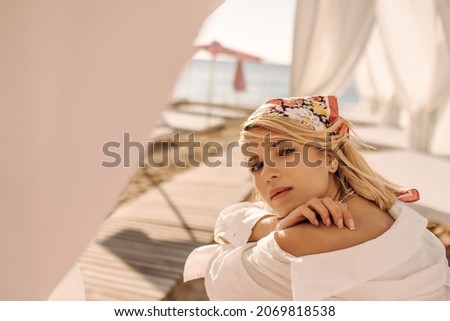 Close-up of young attractive woman sitting near sea and breathing fresh air. Blonde with headscarf in white shirt hugs herself with two hands. Concept of enjoying weekend, vacation