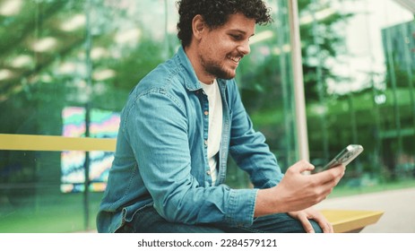 Close-up, young bearded man in denim shirt sits at bus stop waiting for bus and uses mobile phone on modern city background - Powered by Shutterstock