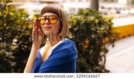 Close-up of young beautiful caucasian girl looking at camera through sunglasses during day on street. Brown-haired woman with bob haircut and bangs wears blue sweatshirt. City life concept Stock photo © 