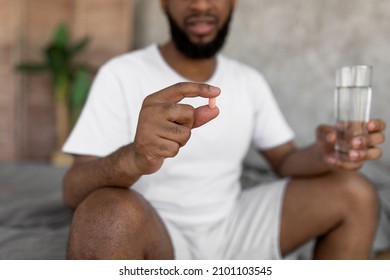 Closeup Of Young Black Man Sitting On Bed With Glass Of Water, Taking Sleeping Pills Or Painkillers At Home. Unrecognizable Millennial Male Using Medications, Holding Tablet In Hand, Selective Focus