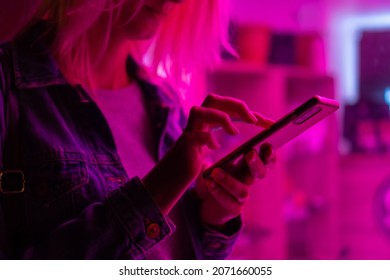 Close-up of a young blonde woman with a mobile phone in her hands in the pink neon light.Pretty young woman in casual street clothes is typing a message on her smartphone.Paying in online banking - Powered by Shutterstock