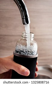 Closeup Of A Young Cacausian Man Filling A Glass Reusable Water Bottle With Water From A Tap Of A Filtered Water Station Or A Domestic Kitchen