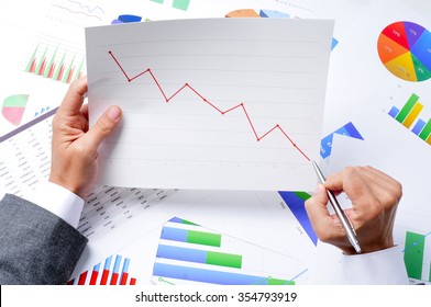 Closeup Of A Young Caucasian Businessman At His Office Desk Full Of Graphs And Charts Observing A Chart With A Downward Trend