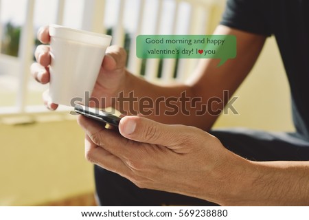 closeup of a young caucasian man with a cup of coffee in his hand sending or reading a text message with a smartphone with the text good morning and happy valentines day, I love you