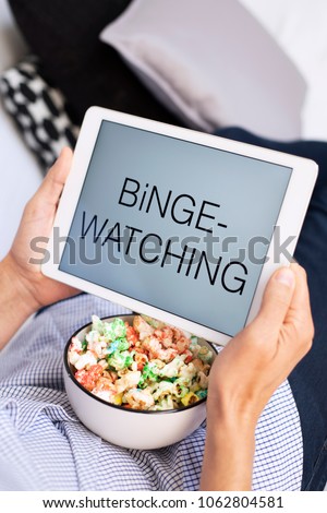 closeup of a young caucasian man lying comfortably on a couch, eating popcorn while is watching his tablet, with the text binge-watching in its screen
