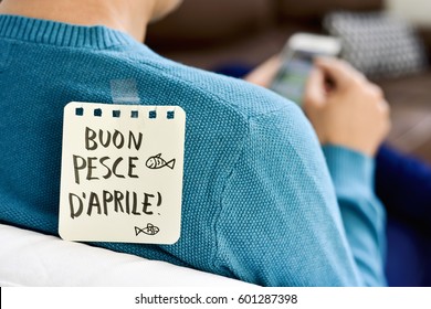 closeup of a young caucasian man seen from behind using his smartphone, with a note with the text buon pesce d aprile, happy april fools day in italian, attached with tape to his back