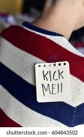 Closeup Of A Young Caucasian Man Seen From Behind Sitting To His Office Desk, With A Note With The Text Kick Me Attached With Tape To His Back