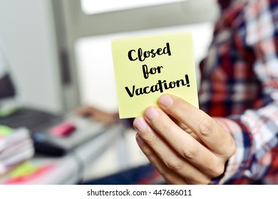 Closeup Of A Young Caucasian Man Showing A Yellow Note With The Text Closed For Vacation, Seating At His Office Desk