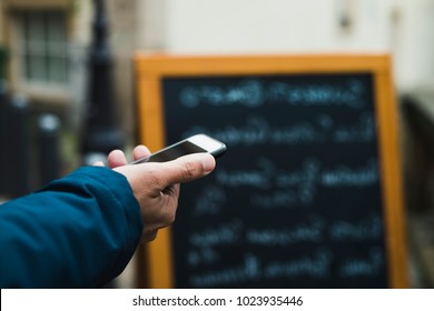 Closeup Of A Young Caucasian Man Using A Smartphone In The Street, In Front Of A Restaurant, Reading The Online Reviews Of The Premises Or Reserving A Table