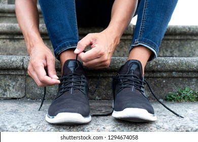 closeup of a young caucasian man, wearing denim pants, sitting on a staircase on the street, tying or untying the shoelaces of his sneakers - Powered by Shutterstock