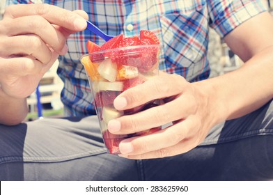 Closeup Of A Young Caucasian Man Wearing A Plaid Shirt Eating A Fruit Salad In A Clear Plastic Cup Outdoors