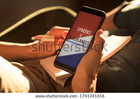 closeup of a young caucasian man, wearing casual clothes, sitting on an armchair, having his smartphone in his hands with the text do you speak Dutch and the flag of the Netherlands in its screen