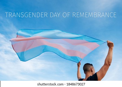 closeup of a young caucasian person, seen from behind, holding a transgender pride flag on the air and the text transgender day of remembrance on the sky