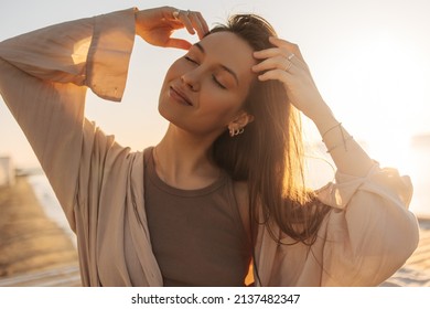 Close-up of young caucasian relaxed woman touching her hair in bright sunlight. Brown-haired girl with closed eyes, wearing brown T-shirt and blouse on top. Good mood, fashion trends