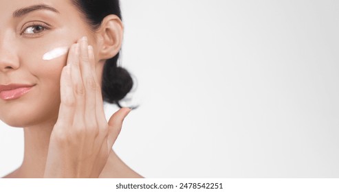 Close-up of a young Caucasian woman applying skincare cream to her face, with ample copy space on a light background. Perfect for beauty, skincare, and wellness advertisement banners. High quality - Powered by Shutterstock