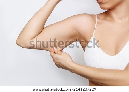 Close-up of a young caucasian woman grabbing skin on her upper arm with excess fat isolated on a white background. Pinching the loose and saggy muscles. Overweight, liposuction concept