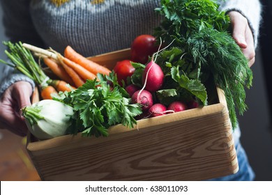 closeup of young Caucasian woman with grey woven sweater holding a large wooden crate full of raw freshly harvested vegetables - Powered by Shutterstock