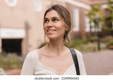 Close-up Of Young Caucasian Woman Looking Seductively Into Side And Smiling, Covering Half Of Face. Beauty With Low Bun Hair, Wears Top And Bag On Shoulder.