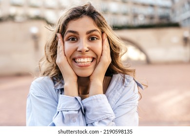 Close-up Of Young Caucasian Woman Pleasantly Surprised Looking At Camera Grabbing Her Face With Hands. Blonde Snow-white Smile, Wears Shirt. Lifestyle, Different Emotions, Leisure Concept.