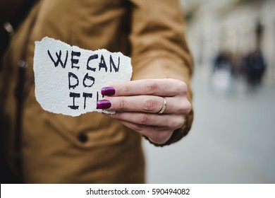closeup of a young caucasian woman in the street showing a piece of paper with the text we can do it written in it