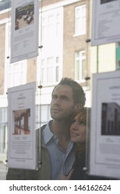 Closeup Of A Young Couple Looking Through Window At Estate Agents