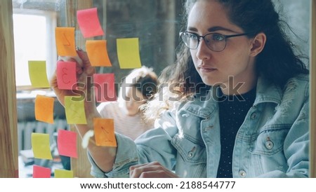 Close-up of young creative entrepreneur numbering coloured stickers on glassboard while working together with female colleague in modern office indoors