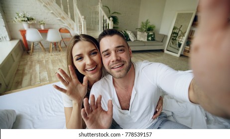 Closeup of Young cute and loving couple having video chat holding smartphone and chatting to friends sitting in bed at home