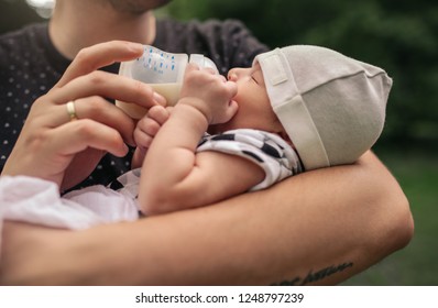 Closeup Of A Young Father Holding His Baby Boy In His Arms And Feeding Him With A Formula Bottle While Standing Outside In A Park