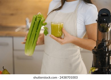 Close-up young female in apron standing in the kitchen. Woman hold glass with green healthy beverage in one hand and celery in another. Detox diet. Homemade juice. Vegetarian. Juicer. - Powered by Shutterstock