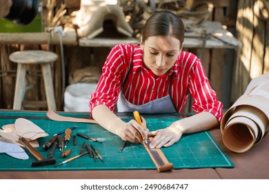 Closeup young female leather maker using a cutter knife to cut a piece of leather sheet according to the design to bring out to sew into products for customers in workshop. - Powered by Shutterstock