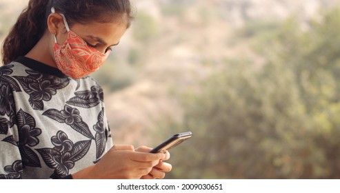 A Closeup Of A Young Female South Asian Child Texting On Her Phone While Wearing An Orange Covid Mask