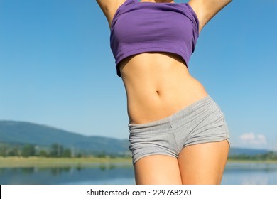 Closeup of young fit woman's belly outdoors in summer. Fitness girl in gray shorts and purple top outdoors at the beach. No retouch.