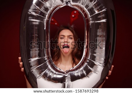 Close-up of young funny pretty female with wavy hairstyle fooling and making faces while posing over claret background with huge air number balloon. Holiday preparation and celebration concept