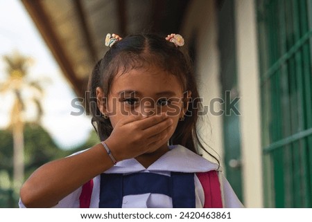 Close-up of a young girl in a school uniform covering her mouth with her hand, standing outside her school.