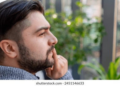 close-up of young guy with a beard thoughtfully looks out the window among the plants