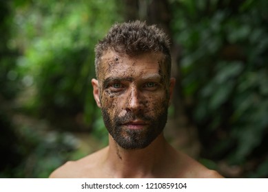 Close-up of a young handsome man with dirty face looks intently at the camera in a wild forest