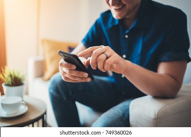 Closeup of young happy smiling man using smartphone device while chilling on sofa a home, cheerful hipster guy typing an sms message, flare light 