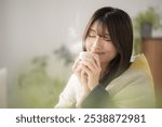 Close-up of a young Japanese (Asian) woman with eyes closed relaxing in a winter living room drinking a hot beverage