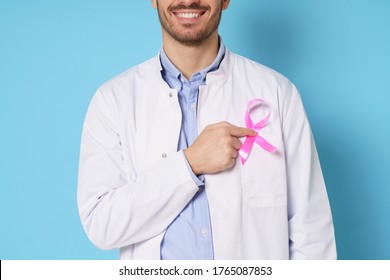 Close-up Of Young Male Oncologist, Dressed In White Medical Robe, Pointing At Pink Ribbon, Raising Awareness Of Breast Cancer, Smiling, Isolated On Blue Background