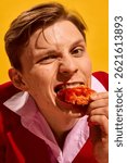 Close-up of young man biting into spicy chicken wing with intense expression against bold yellow background. Concept of fast food aesthetics, pop art photography, emotions