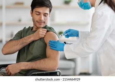 Closeup of young man in camouflage uniform soldier in wheelchair getting immunization against COVID-19 at clinic, unrecognizable nurse in protective workwear making shot in shoulder for military man - Powered by Shutterstock