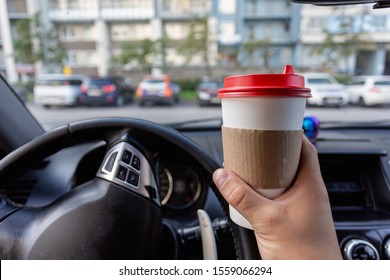 Closeup of a young man car driver drinking coffee, hand holding a paper white coffee Cup with a red lid in the background steering car dashboard blurred Parking background.