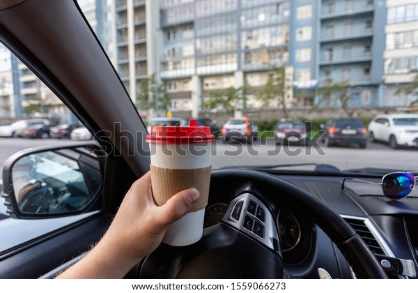 Closeup of a young
man car driver drinking coffee, hand holding a paper white coffee
Cup with a red lid in the background steering car dashboard blurred
Parking background.