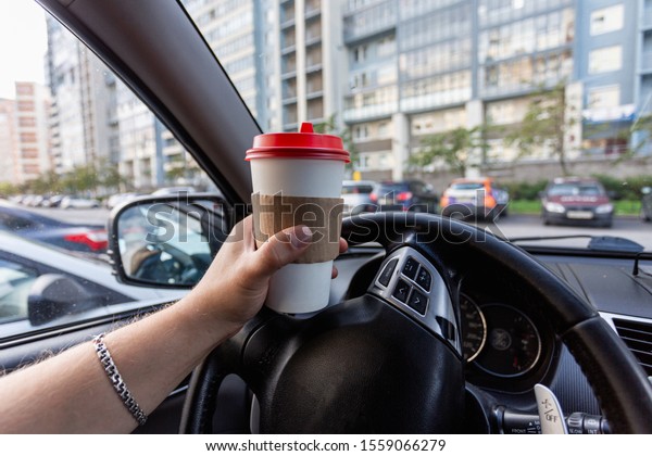 Closeup of a young\
man car driver drinking coffee, hand holding a paper white coffee\
Cup with a red lid in the background steering car dashboard blurred\
Parking background.