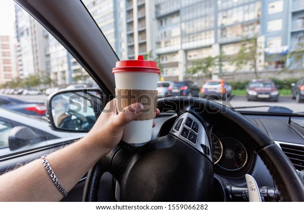 Closeup of a young\
man car driver drinking coffee, hand holding a paper white coffee\
Cup with a red lid in the background steering car dashboard blurred\
Parking background.