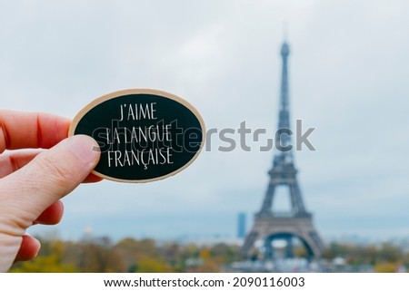 closeup of a young man holding a black signboard with the text I love French language written in French in it, in front of the Eiffel Tower in Paris, France