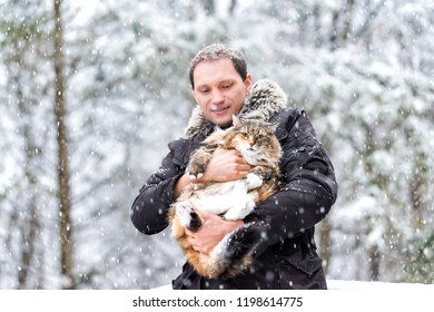 Closeup Of Young Man Holding Curious Maine Coon Cat Outside, Outdoors In Park In Snow, Snowing Weather During Snowstorm, Storm With Snowflakes, Flakes Falling